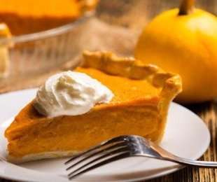 Na zdjęciu talerzyk, na nim porcja ciasta dyniowego z bitą śmietaną, w tle stoi dynia