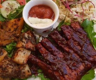 Na zdjęciu talerz, na nim potrawa Żeberka z Tempehu w sosie barbecue podawane z pieczonymi ziemniakami, warzywami i sosami